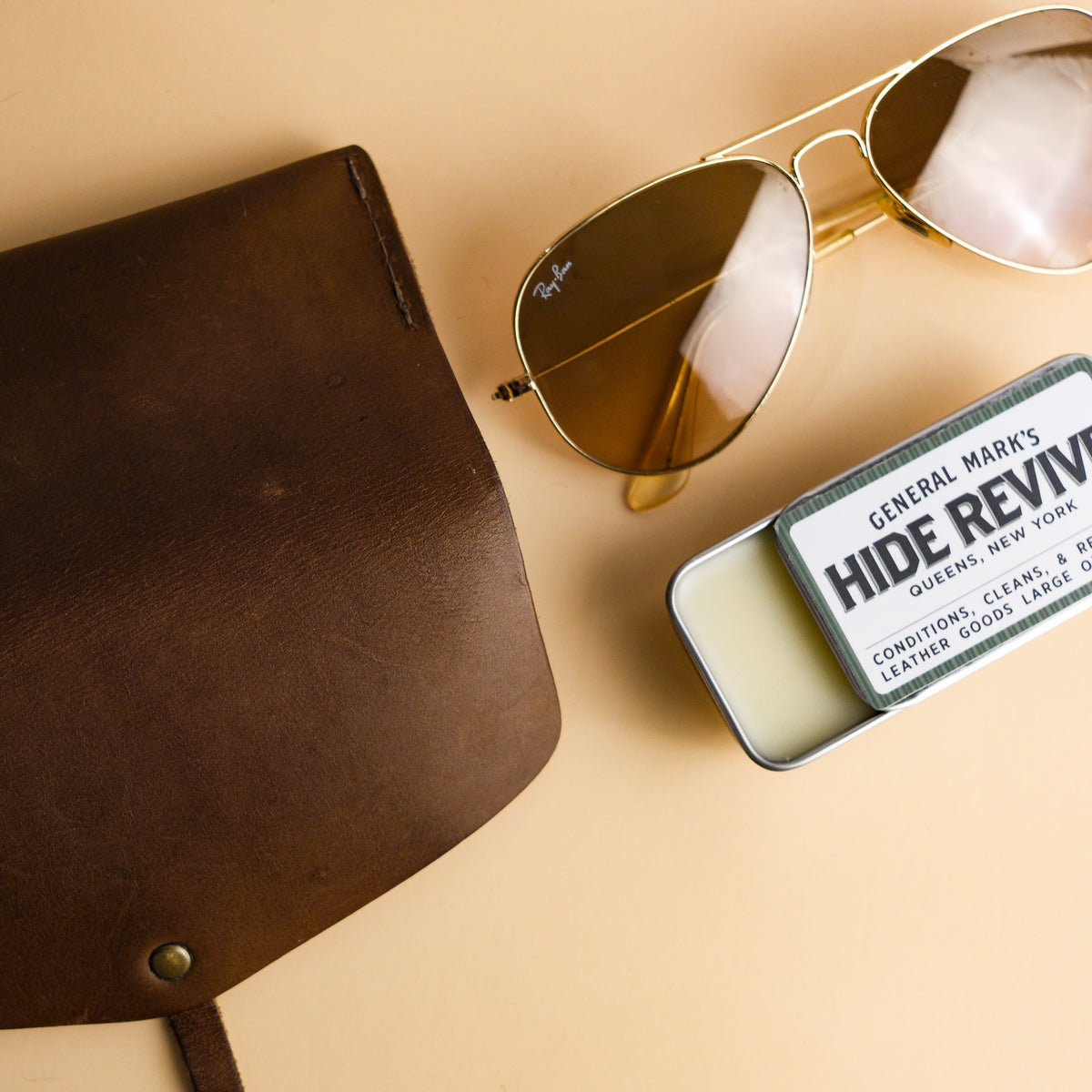 Open tin of General Mark’s Hide Revive natural leather conditioner beside a brown leather case and aviator sunglasses